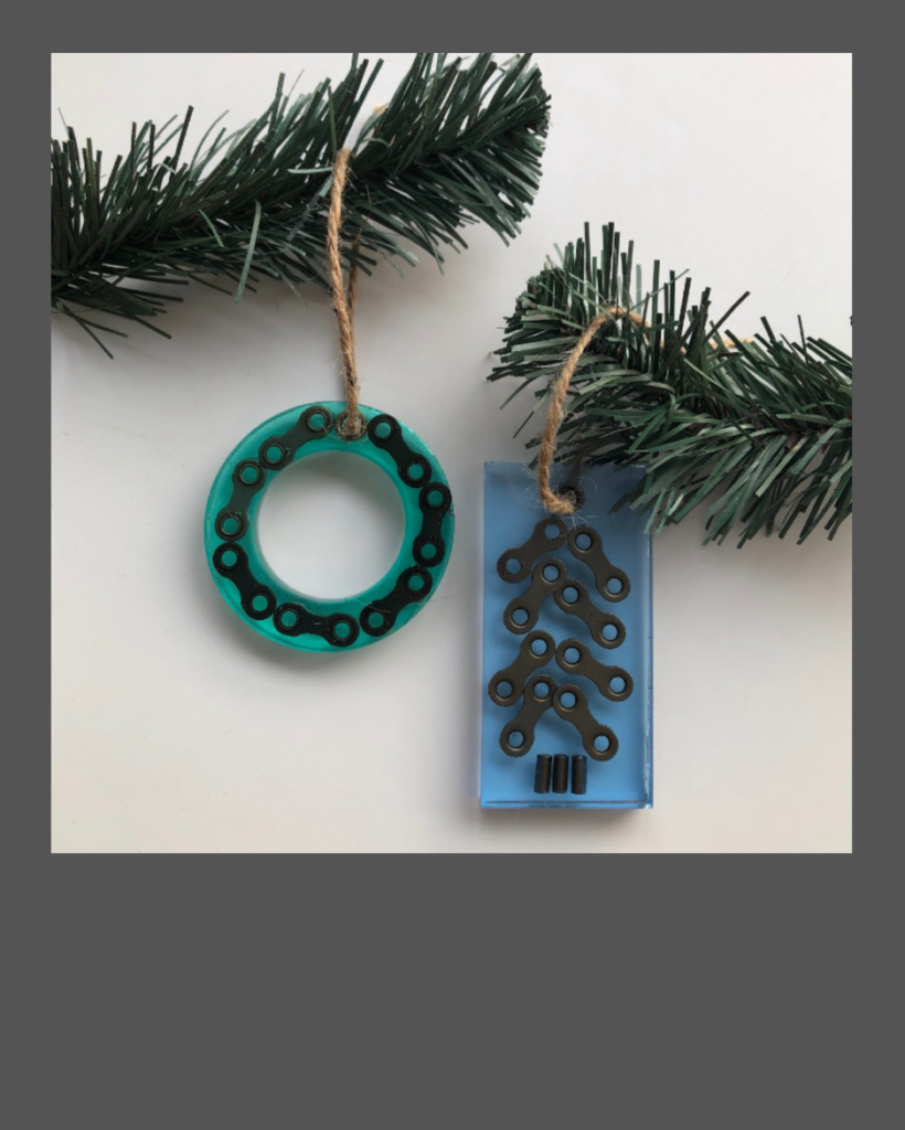Two Christmas ornaments made of blue and green epoxy resin containing pieces of bicycle chain hanging from evergreen branches.